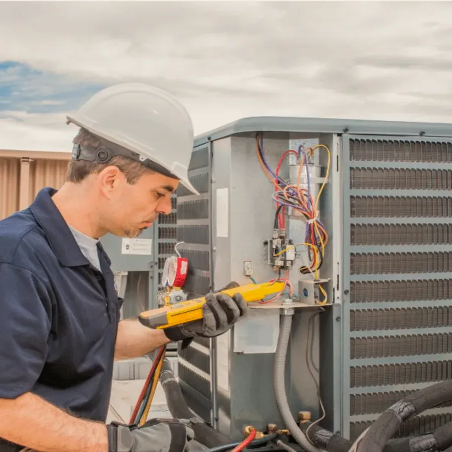 HVAC technician in a hard hat testing an outdoor air conditioner with a multimeter
